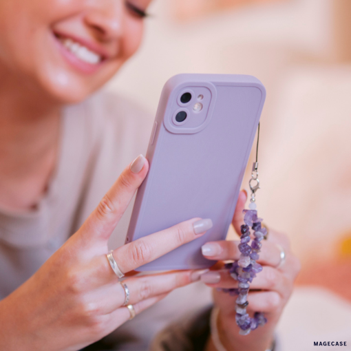A person with light pink nail polish from the NAKD spring collection smiles while holding a smartphone adorned with the "NAKD Phone Case - Lilac" by Coconut Lane, which offers Full Camera Protection and features an amethyst crystal charm. The background is softly blurred, highlighting the person's joyful expression and the phone accessories.