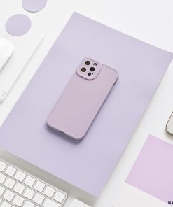 A lilac-colored smartphone case from the Coconut Lane NAKD collection is placed on a matching lilac sheet of paper. Surrounding it are various office supplies, including a white keyboard, a white pencil, a white wireless earbud, and a partially visible laptop. The setting is minimalistic and clean.
