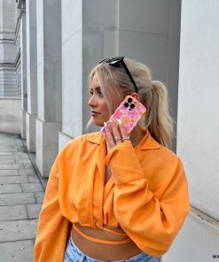 A person with blonde hair, wearing black sunglasses on their head, an orange button-up shirt, and blue jeans, is holding a smartphone with a Coconut Lane Wavy Phone Case - Pink & Orange Bloom to their ear. They are standing on a street with light-colored buildings in the background.