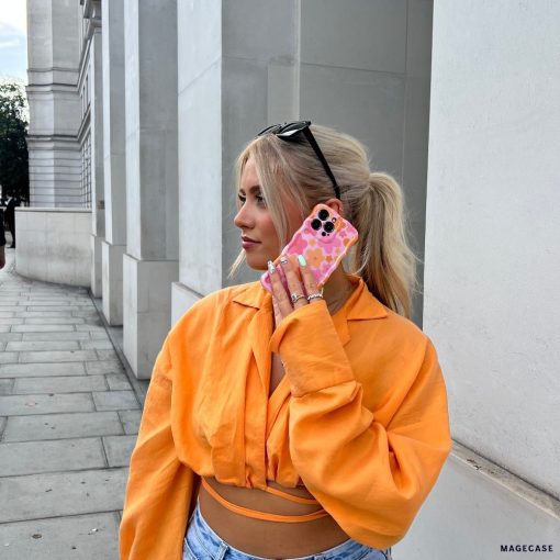 A person with blonde hair, wearing black sunglasses on their head, an orange button-up shirt, and blue jeans, is holding a smartphone with a Coconut Lane Wavy Phone Case - Pink & Orange Bloom to their ear. They are standing on a street with light-colored buildings in the background.