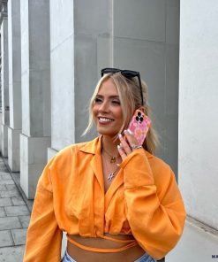 A woman with long blonde hair, wearing a bright orange cropped shirt and light blue jeans, stands on a sidewalk smiling while holding a phone with an extra protective Wavy Phone Case - Pink & Orange Bloom by Coconut Lane to her ear. She has sunglasses perched on her head and a necklace with a capital letter 