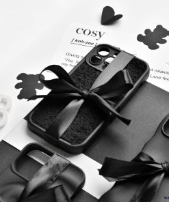 A monochrome image featuring the Cosy Teddy Phone Case - Black by Coconut Lane, tied with a black ribbon and placed on a black-and-white textured surface. Also shown are a dictionary definition of 