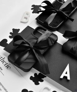 A monochrome photo featuring Coconut Lane's Melting Heart Phone Cases in Matte Black, elegantly tied with black ribbons. They are arranged on a black and white surface adorned with scattered letters and cut-out shapes, with the word 