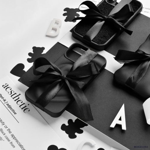 A monochrome photo featuring Coconut Lane's Melting Heart Phone Cases in Matte Black, elegantly tied with black ribbons. They are arranged on a black and white surface adorned with scattered letters and cut-out shapes, with the word "aesthetic" partially visible on a sheet of paper in the background.