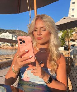A blonde woman, seated at an outdoor café by the waterfront, holds a Coconut Lane 3D Gummy Phone Case - Pink shaped like a bear. Dressed in a chic and colorful floral outfit, she accessorizes with jewelry and an Apple Watch. The background features buildings, hills, and umbrellas under a clear blue sky.