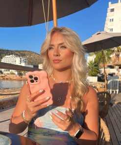 A woman with long blonde hair sits outside at a wooden table under an umbrella, holding a 3D Gummy Phone Case - Pink from Coconut Lane shaped like a bear. She is dressed in a light blue dress, has several rings on her fingers and painted nails. The background features buildings and palm trees.