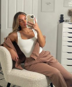 A young woman with long blonde hair is taking a mirror selfie with her iPhone, which is covered by the Cream Curvy Phone Case from Coconut Lane, while sitting in a white boucle chair. She is wearing a white tank top and brown pants, with a brown jacket draped over her shoulders. Behind her, there are framed pictures on a white wall and a white dresser.