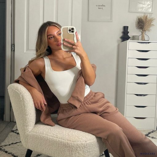 A young woman with long blonde hair is taking a mirror selfie with her iPhone, which is covered by the Cream Curvy Phone Case from Coconut Lane, while sitting in a white boucle chair. She is wearing a white tank top and brown pants, with a brown jacket draped over her shoulders. Behind her, there are framed pictures on a white wall and a white dresser.