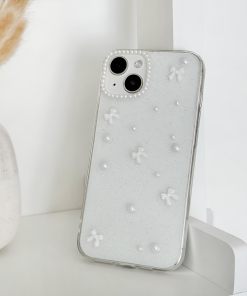 A white smartphone with a Mini White Bows Phone Case by Coconut Lane, featuring small 3D flowers and pearl-like accents, is propped up against a white wall. In the background, a portion of a white vase with dried beige grass sticking out adds to the minimalist and monochromatic setting.