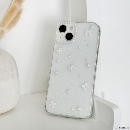 A white smartphone with a Mini White Bows Phone Case by Coconut Lane, featuring small 3D flowers and pearl-like accents, is propped up against a white wall. In the background, a portion of a white vase with dried beige grass sticking out adds to the minimalist and monochromatic setting.