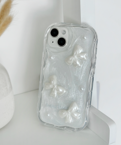 A **Coconut Lane** Pearls & Bows Phone Case, adorned with white textured bows and pearl-like accents, rests on a white shelf. Nearby, a white vase filled with dried wheat stalks adds a decorative touch to the background.