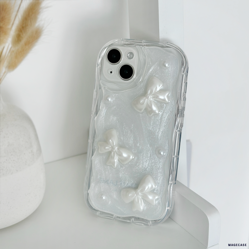 A **Coconut Lane** Pearls & Bows Phone Case, adorned with white textured bows and pearl-like accents, rests on a white shelf. Nearby, a white vase filled with dried wheat stalks adds a decorative touch to the background.