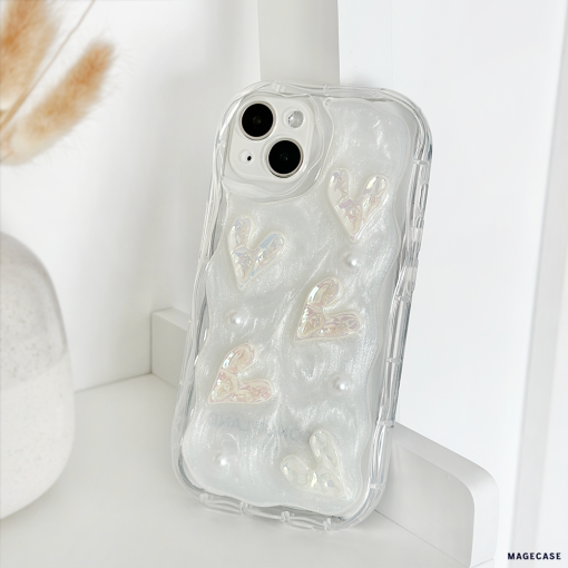 The Pearly Hearts Phone Case - Clear by Coconut Lane is an iridescent phone case with a textured design featuring raised, shimmering hearts and small pearl-like embellishments. This extra protective case is propped up on a white surface next to a vase containing dried wheat stalks.