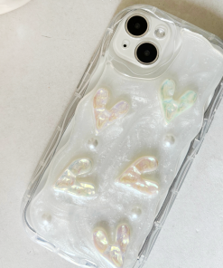 A close-up image of an iPhone with a Coconut Lane Pearly Hearts Phone Case - Clear. The case, clear with a pearl white shimmer effect, is adorned with raised iridescent heart shapes and small pearl-like beads. Extra protective, the phone is placed on a light gray surface.