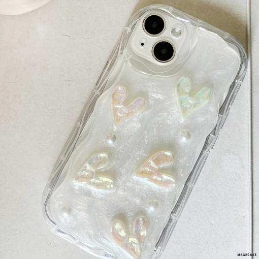 A close-up image of an iPhone with a Coconut Lane Pearly Hearts Phone Case - Clear. The case, clear with a pearl white shimmer effect, is adorned with raised iridescent heart shapes and small pearl-like beads. Extra protective, the phone is placed on a light gray surface.