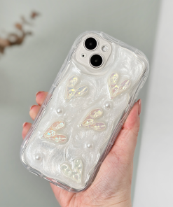 A hand holds an iPhone15 with an extra Coconut Lane Pearly Hearts Phone Case - Clear. The case showcases a pearlescent white design with raised heart shapes and small pearl-like embellishments. The phone is held in front of a blurred background.