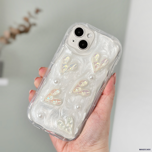 A hand holds an iPhone15 with an extra Coconut Lane Pearly Hearts Phone Case - Clear. The case showcases a pearlescent white design with raised heart shapes and small pearl-like embellishments. The phone is held in front of a blurred background.