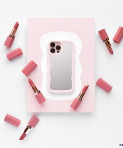 A phone with the Curvy Mirror Phone Case in pink from Coconut Lane lies on a pink and white background, surrounded by eight pink lipstick tubes, some with their caps off, revealing the lipsticks. The composition is minimalistic with a soft color palette.