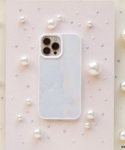 A white smartphone encased in a Coconut Lane Mother of Pearl Phone Case is centered on a light, glittery surface. The phone is surrounded by variously sized pearls scattered around, creating a delicate and elegant visual composition.
