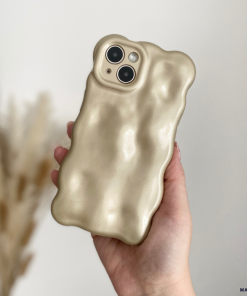A person is holding a beige-colored iPhone 15 with Coconut Lane's distinctive 3D Pearl Phone Case - Gold. The softly blurred background features dried pampas grass, highlighting the soft and cushiony texture of the gold-accented case.