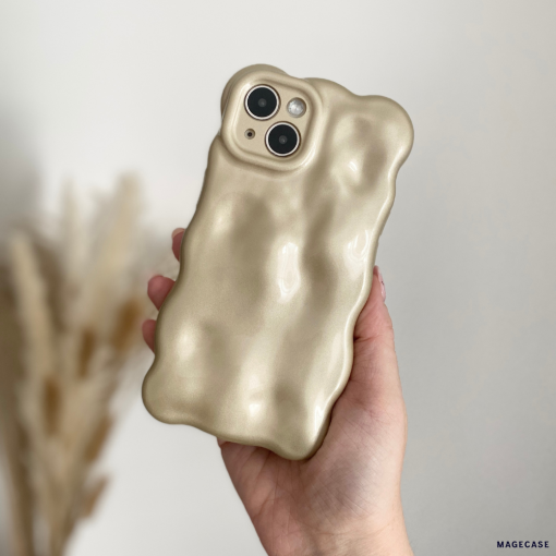 A person is holding a beige-colored iPhone 15 with Coconut Lane's distinctive 3D Pearl Phone Case - Gold. The softly blurred background features dried pampas grass, highlighting the soft and cushiony texture of the gold-accented case.