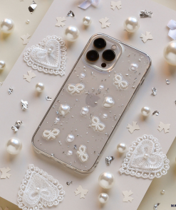 The Coquette Dreams Phone Case by Coconut Lane, crafted as an ultra-protective iPhone 15 case, features a clear base adorned with white lace patterns, pearls, and glitter. It is displayed on a decorated surface that includes scattered white lace hearts, tiny pearls, metallic star-shaped confetti, and small white bows.
