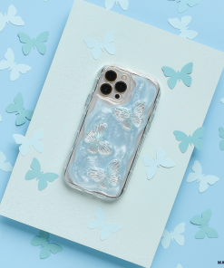 A smartphone with the Coconut Lane Shimmer Blue Butterflies Phone Case rests on a textured blue surface, surrounded by various paper cutouts of butterflies in shades of blue and white. The phone's camera lenses are visible at the top.