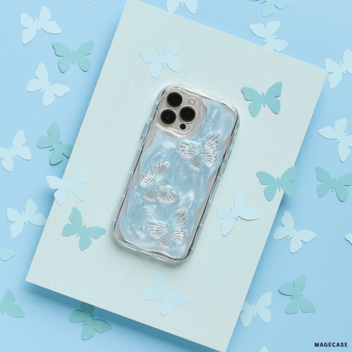 A smartphone with the Coconut Lane Shimmer Blue Butterflies Phone Case rests on a textured blue surface, surrounded by various paper cutouts of butterflies in shades of blue and white. The phone's camera lenses are visible at the top.