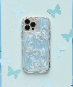 A silver smartphone fitted with Coconut Lane's Shimmer Blue Butterflies Phone Case, adorned with three iridescent butterfly designs, is surrounded by cut-out paper butterflies on a pastel blue and turquoise background. The phone features a triple camera on the top left corner.
