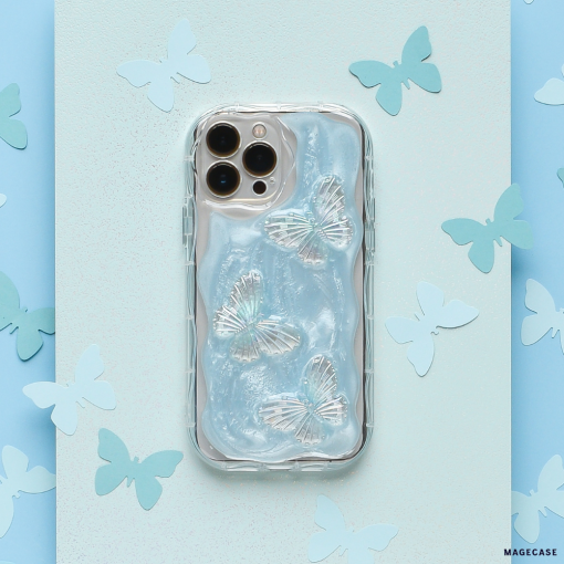 A silver smartphone fitted with Coconut Lane's Shimmer Blue Butterflies Phone Case, adorned with three iridescent butterfly designs, is surrounded by cut-out paper butterflies on a pastel blue and turquoise background. The phone features a triple camera on the top left corner.