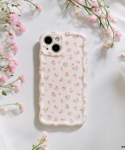 A smartphone with the Wavy Phone Case – Ditsy Floral Pink by Coconut Lane lies on white fabric. Small pink flowers encircle the phone, enhancing the scene with a soft, elegant touch.