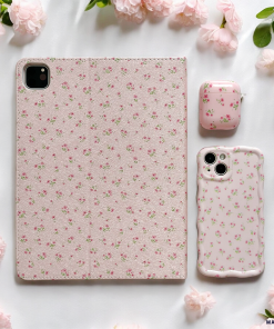 A tablet, a phone protected by Coconut Lane's Wavy Phone Case in Ditsy Floral Pink, and an AirPods case all featuring matching pink floral designs are arranged on a white surface surrounded by pale pink blossoms. The cases showcase a delicate pattern of ditsy floral pink flowers, creating a cohesive and aesthetically pleasing look.