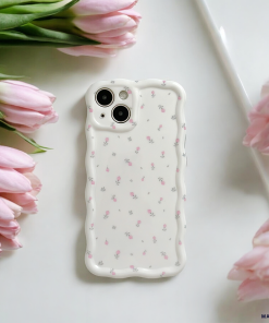 A white smartphone adorned with the Wavy Phone Case - Ditsy Floral White by Coconut Lane, featuring a dainty, scalloped edge and decorated with small pink flowers, is placed on a white surface surrounded by pink tulips. The arrangement exudes a soft and elegant aesthetic.