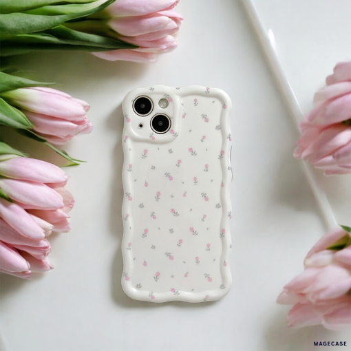 A white smartphone adorned with the Wavy Phone Case - Ditsy Floral White by Coconut Lane, featuring a dainty, scalloped edge and decorated with small pink flowers, is placed on a white surface surrounded by pink tulips. The arrangement exudes a soft and elegant aesthetic.