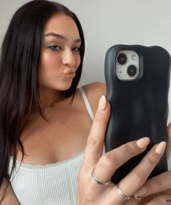A woman with long dark hair takes a selfie with her smartphone, showcasing the 3D Boujee Phone Case - Matte Black by Coconut Lane. She wears a white tank top, makes a pouty face, and holds the phone in her left hand against a plain white background.