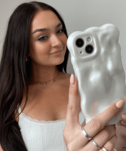 A young woman with long brown hair holds up her iPhone15 encased in Coconut Lane's 3D Pearl Phone Case - White. She is smiling and wearing a white tank top with lace trim, along with several rings and a delicate necklace. The background is plain and light-colored, emphasizing the pearl white effect of her phone case.