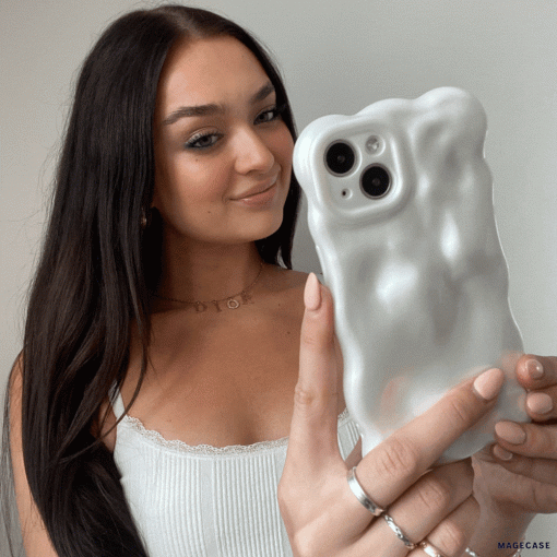 A young woman with long brown hair holds up her iPhone15 encased in Coconut Lane's 3D Pearl Phone Case - White. She is smiling and wearing a white tank top with lace trim, along with several rings and a delicate necklace. The background is plain and light-colored, emphasizing the pearl white effect of her phone case.