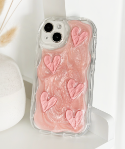 A Coconut Lane Shimmer Pink Hearts Phone Case featuring an extra protective clear design with pink swirls and five raised shimmer pink hearts is propped up against a white vase with dried wheat stalks. The background is a clean, white surface.
