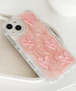 A smartphone encased in Coconut Lane's Shimmer Pink Hearts Phone Case, an extra protective transparent cover showcasing a pink and white marbled design adorned with six raised shimmer pink hearts. The phone is set on a light-colored speckled surface, creating a clean and minimalist aesthetic.